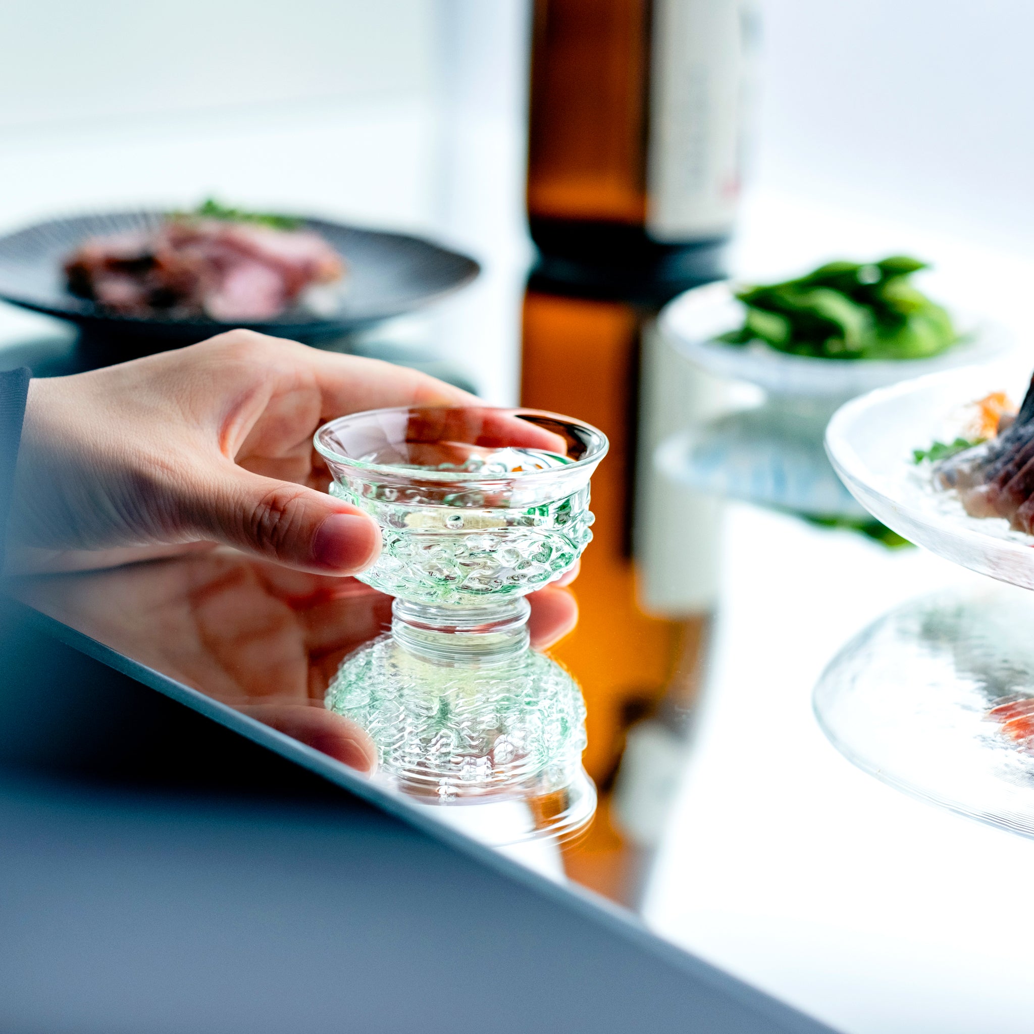 津軽びいどろ 盃12ヶ月コレクション 5月新緑 食卓に並ぶ盃を手でつかんでいる画像