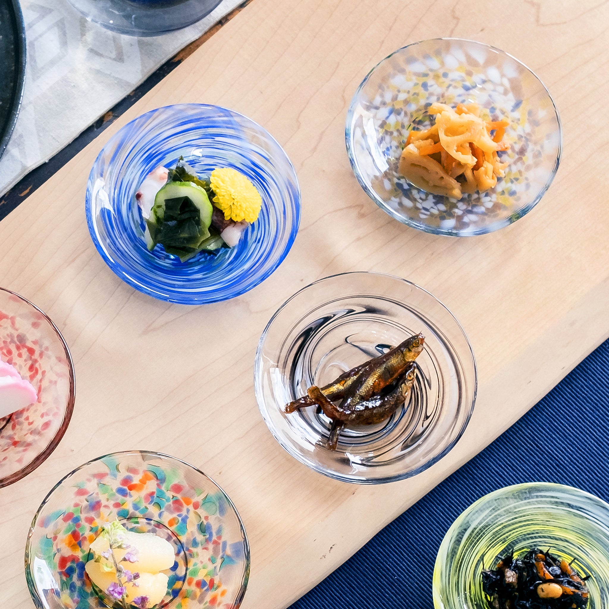 津軽びいどろ 色色豆皿 夜雲 料理が乗った色違いの小皿がテーブルに並んでいる画像