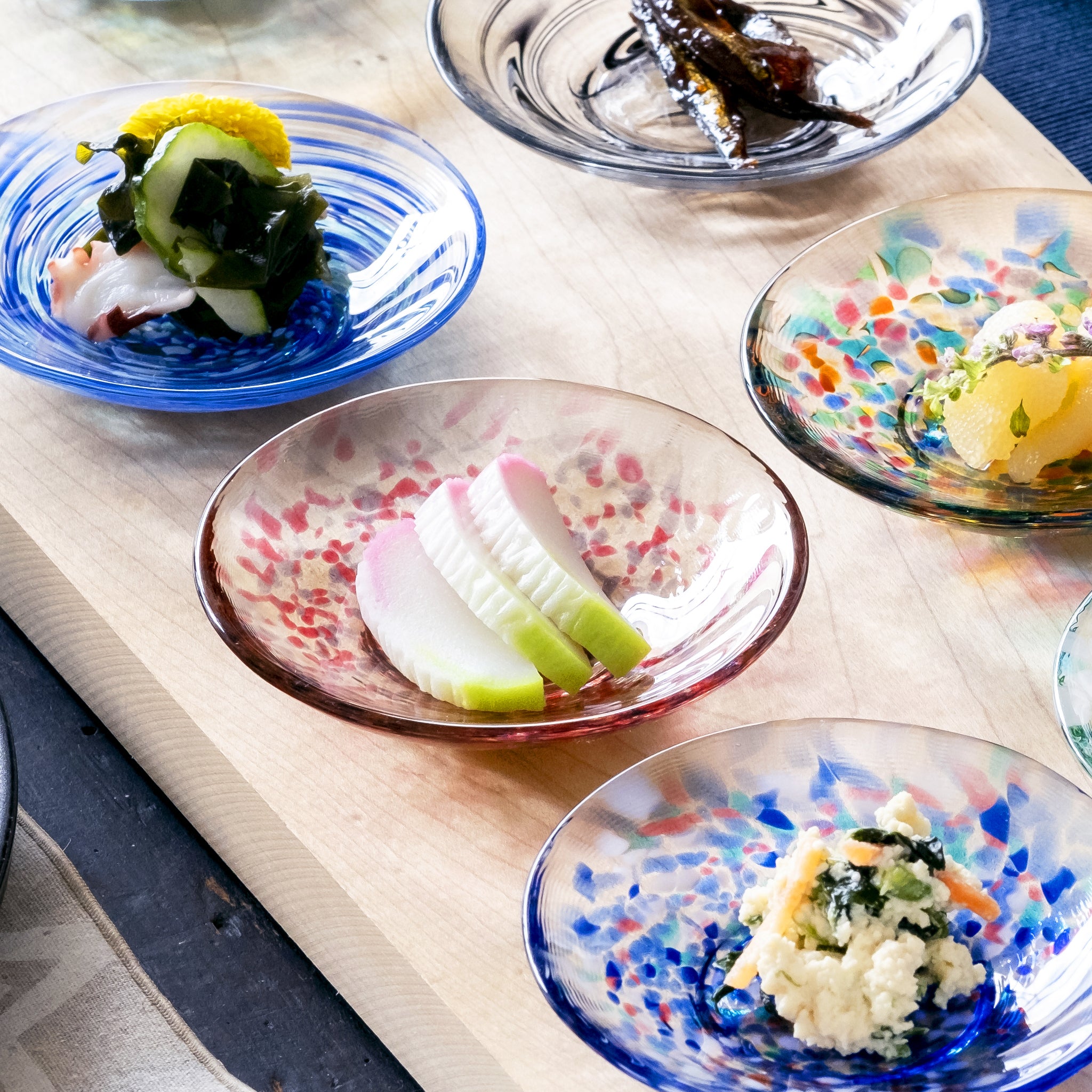 津軽びいどろ 色色豆皿 春茜 料理が乗った小皿がテーブルに並んでいる画像