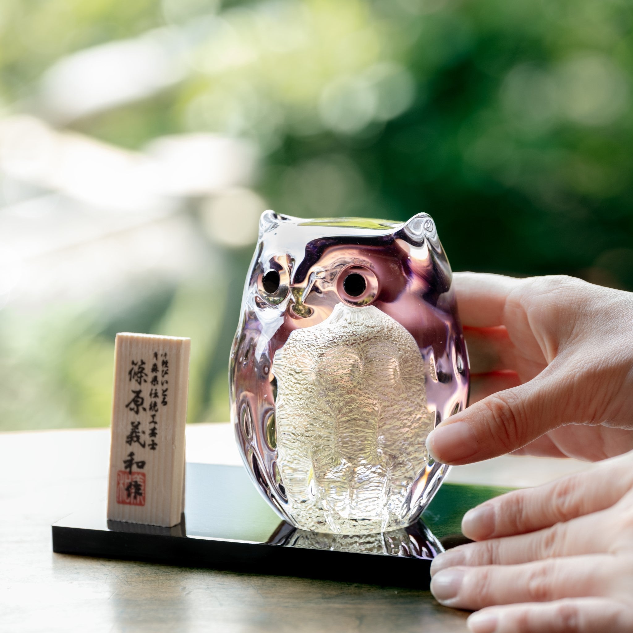 津軽びいどろ 親ふくろう (紫暮) 紫色のフクロウの置物に手を添えている画像